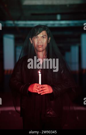 un adoratore del diavolo maschio con un velo trasparente sta eseguendo un rituale spettrale tenendo una candela in mano la notte Foto Stock