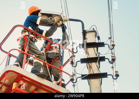 Due elettricisti dalla base della piattaforma aerea o della gru stanno riparando la lampada per illuminazione stradale. Elettricisti professionisti che indossano caschi, tute e assicurazioni in altezza. Vista dei lavoratori dal basso verso il cielo. Flusso di lavoro autentico. Foto Stock