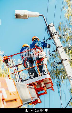 Due elettricisti professionisti con elmetti stanno riparando le linee elettriche dalla base del dumper a benna. Vista dal basso. Gli elettricisti cambiano i cavi sui pali per illuminazione stradale. Foto Stock