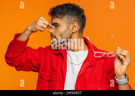 Stanco e stanco giovane indiano si toglie gli occhiali, sente dolore agli occhi, essendo il lavoro eccessivo bruciato a causa delle lunghe ore di lavoro. Assonnato ragazzo arabo esausto che si sfrega gli occhi isolato su sfondo arancione in interni Foto Stock