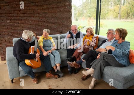 In una sala accogliente e ben illuminata con una grande finestra che offre una vista sull'esterno, un gruppo di anziani condivide un momento di divertimento musicale. Uno di loro sta suonando una classica chitarra acustica, mentre gli altri ascoltano attentamente, alcuni con gli occhi chiusi, battendo i piedi e mostrando espressioni di appagamento e relax. L'ambiente confortevole e l'esperienza musicale condivisa creano un'atmosfera di calore e di amicizia. Melodie musicali tra amici, anziani che si godono una sessione di chitarra. Foto di alta qualità Foto Stock