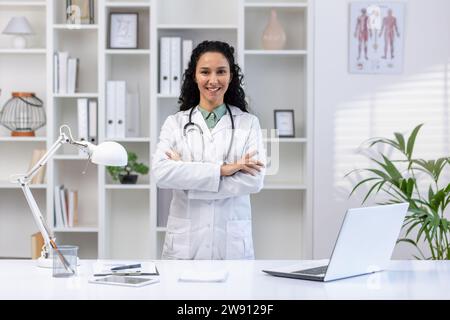 Ritratto di giovane bella medico di successo all'interno dello studio medico, donna ispanica con i capelli ricci in cappotto medico bianco sorridente e guardando la macchina fotografica con le braccia incrociate, medico all'interno della clinica. Foto Stock