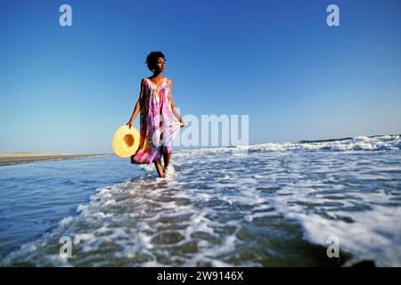Una donna che si prende cura di un cappello di paglia che cammina da sola attraverso il surf in arrivo su una spiaggia deserta nel North Carolina Foto Stock