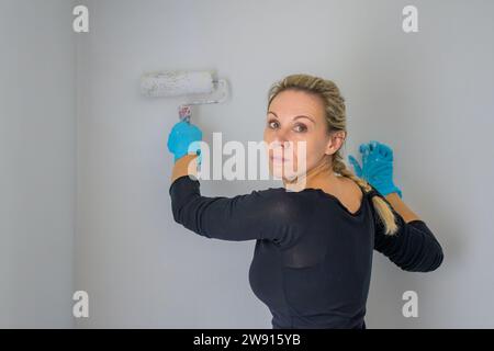 Bionda donna single che si gira e guarda la macchina fotografica mentre dipinge il suo appartamento con un grande rullo di vernice nel suo nuovo appartamento Foto Stock