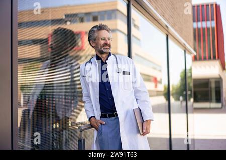 Medico sorridente in piedi con un computer portatile vicino al vetro nelle giornate di sole Foto Stock