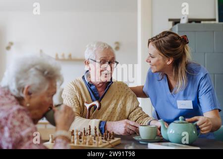 Lavoratore sanitario felice che serve tè alla coppia senior a tavola Foto Stock