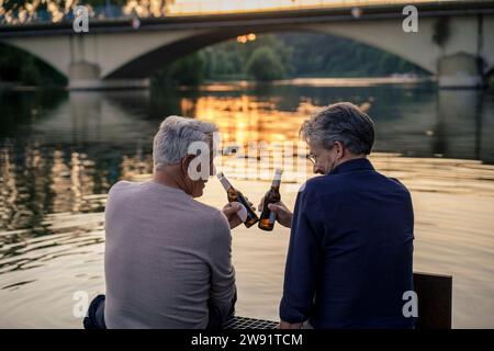 Amici anziani che tostano bottiglie di birra vicino all'acqua al tramonto Foto Stock