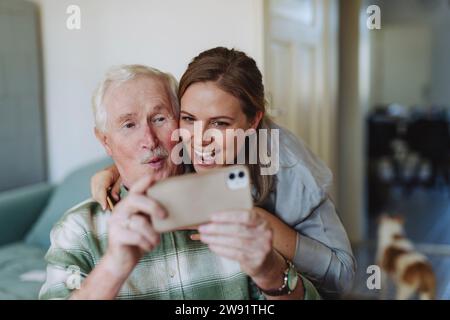 Lavoratore sanitario felice con uomo anziano che prende selfie a casa Foto Stock