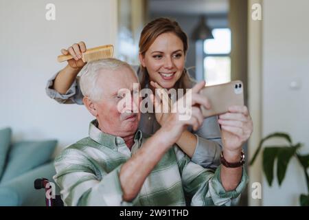 Lavoratore sanitario felice pettinando i capelli dell'uomo anziano prendendo selfie a casa Foto Stock
