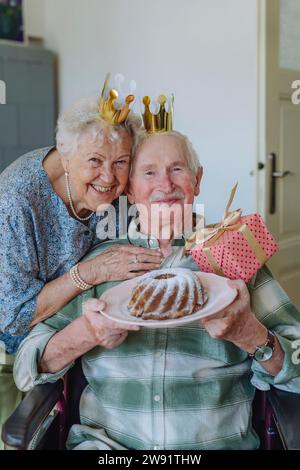 Donna anziana sorridente con un uomo che tiene la torta di compleanno a casa Foto Stock