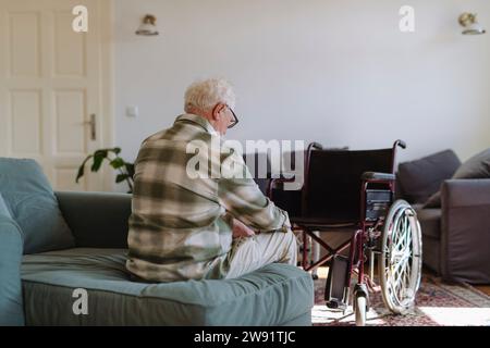 Un uomo anziano triste seduto sul divano vicino alla sedia a rotelle a casa Foto Stock