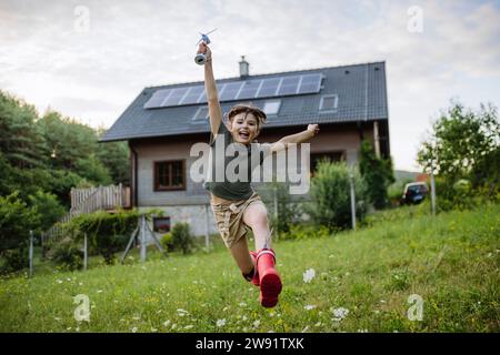 Una bambina che corre davanti alla casa di famiglia con pannelli solari sul tetto che porta il modello a turbina eolica Foto Stock