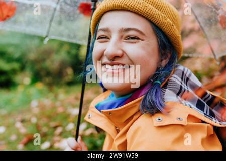 Donna sorridente che indossa un cappello in maglia e tiene un ombrello nel parco autunnale Foto Stock