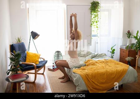 Giovane donna transgender che si allunga con le braccia alzate seduta sul letto a casa Foto Stock