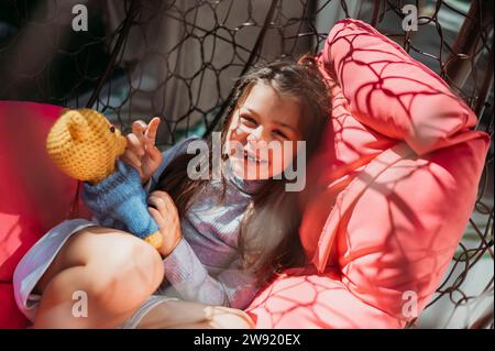 Ragazza sorridente con giocattolo imbottito sdraiata sull'altalena del portico Foto Stock