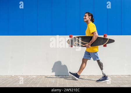 Un giovane felice che tiene lo skateboard e cammina con una gamba protesica davanti al muro Foto Stock