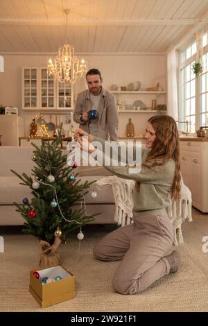 Donna sorridente che decora l'albero di Natale con un uomo in piedi a casa Foto Stock