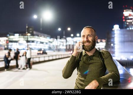 Uomo sorridente che parla con uno smartphone vicino alla ringhiera in città Foto Stock