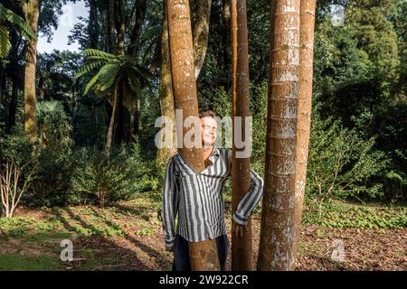 Giovane sorridente in piedi con un albero all'interno della camicia a righe nella foresta Foto Stock