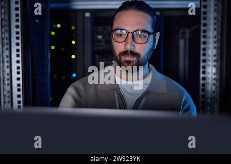Giovane ingegnere che indossa occhiali da vista e lavora su un computer portatile nella sala server Foto Stock
