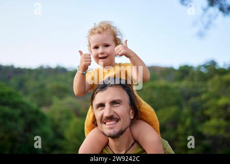 Ragazza carina che mostra i pollici in alto seduta sulla spalla del padre Foto Stock