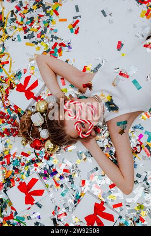 Ragazza sorridente sdraiata con i coriandoli vicino alle confezioni regalo sul pavimento bianco Foto Stock