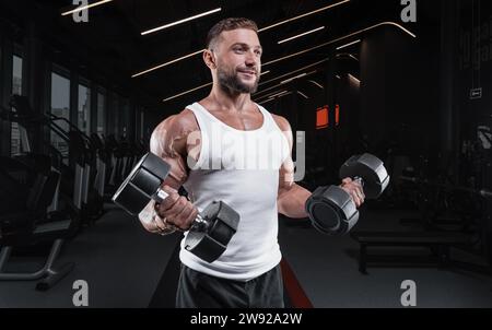 Immagine di un ragazzo muscoloso in una maglia bianca con attrezzature sportive. Palestra. Concetto di fitness Foto Stock