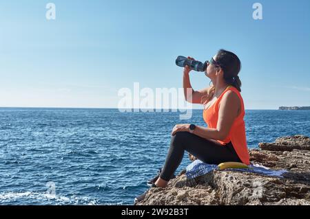 Donna latina, mezza età, riposa, riacquista forza, mangia, acqua potabile, dopo una sessione in palestra, calorie brucianti, mantenersi in forma, all'aperto vicino al Foto Stock