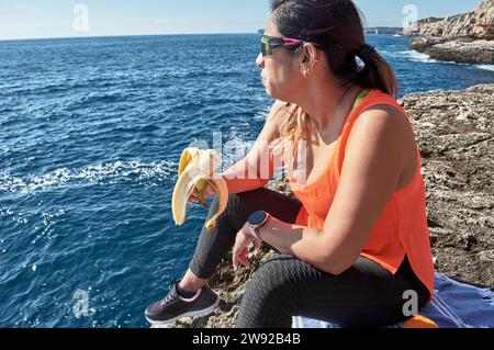 Donna latina, mezza età, riposa, riacquista forza, mangia, acqua potabile, dopo una sessione in palestra, calorie brucianti, mantenersi in forma, all'aperto vicino al Foto Stock