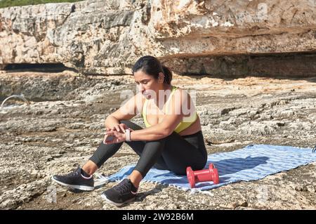 Donna latina, mezza età, guarda app, orologio intelligente, riposa dopo una sessione in palestra, indossa il top giallo, leggings neri, manichino, calorie brucianti Foto Stock