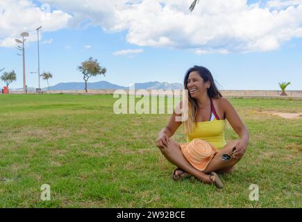 Bella donna latina di 40 anni, sorridente seduta sull'erba di un parco di Maiorca, isole baleari, concetto di vacanza Foto Stock