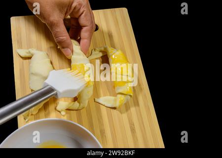 Preparazione di croissant, ripieni di prosciutto e formaggio e dipinti con tuorlo d'uovo, su una tavola di legno, pronti per la cottura Foto Stock