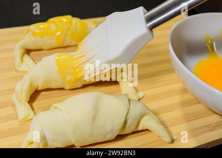 Preparazione di croissant, ripieni di prosciutto e formaggio e dipinti con tuorlo d'uovo, su una tavola di legno, pronti per la cottura Foto Stock