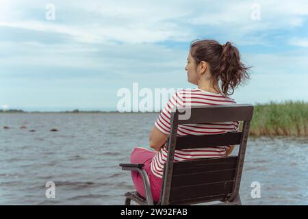 una donna di mezza età siede tranquillamente su una sedia vicino alla spiaggia estiva, assorbendo la tranquillità dell'ambiente circostante. Foto Stock