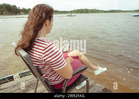 Una donna di mezza età con capelli lunghi e fluenti è seduta sulla riva di un lago tranquillo in una giornata di sole, immersa nel suo smartphone, mentre le piace Foto Stock