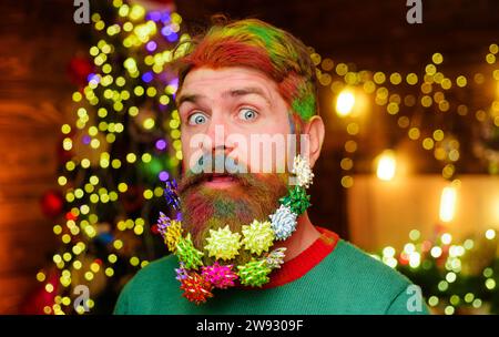 Vacanze invernali. Festa di Natale o di Capodanno. Un uomo barbuto sorpreso con barba decorata e capelli colorati tinti su sfondo natalizio. Barbiere Foto Stock