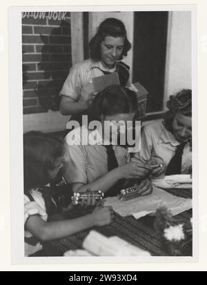 Girls of the Youth Storm, 1942 - 1943 Fotografia quattro ragazze della tempesta giovanile, in uniforme, stanno preparando pacchetti da inviare ai volontari delle SS olandesi sul fronte orientale. Questo è stato fatto attraverso l'organizzazione Frontzorg fondata da Mussert nel 1942. Qui sono confezionati panini, menta piperita e altre caramelle. Sullo sfondo un poster dell'NSB della primavera 1942: Gossiping Your People Shakes. Progettista: Van Altena. Supporto fotografico Paesi Bassi stampa in argento gelatina invio, imballaggio di materiale postale Paesi Bassi Foto Stock