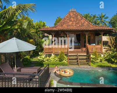 Vista aerea di una giovane donna che fa colazione su un cesto galleggiante in una piscina, nella zona rurale di Bali Foto Stock