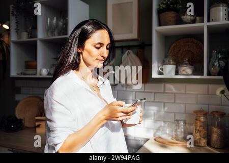 Una giovane donna affascinante tiene uno smartphone e beve un caffè la mattina in cucina. Un'attraente bruna legge le notizie e fa acquisti Foto Stock