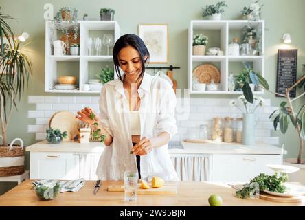 Una giovane donna attraente prepara una deliziosa limonata fresca e annaffia limone e menta. Un'incantevole bruna gode dell'odore degli agrumi e prepara un healt Foto Stock