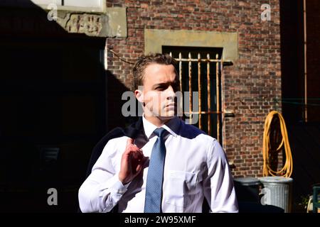 Giovane uomo bianco muscoloso che porta la giacca nera sulla spalla e indossa una camicia bianca con colletto e cravatta blu davanti a una parete di mattoni Foto Stock