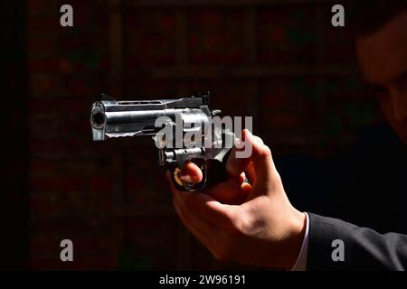 Primo piano di un revolver cromato con un dito sul grilletto di un uomo bianco che indossa una tuta nera Foto Stock