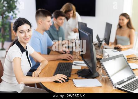 Giovane ragazza entusiasta che studia in un laboratorio informatico con colleghi studenti impegnati in corsi. Concetto di istruzione digitale.. Foto Stock