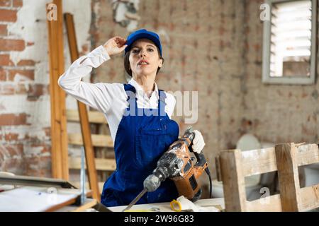 Abile costruttore femminile in tute blu che regge con sicurezza il martello pneumatico mentre si trova all'interno della casa in costruzione Foto Stock