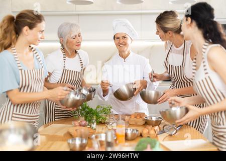 Donna anziana cuoca in abito bianco tiene la ciotola in mano e racconta alle studentesse di corsi culinari su piatti semplici e sani della cucina straniera Foto Stock