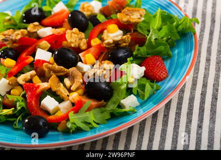 Insalata vitaminica di rucola con pomodori, peperone rosso, feta, fragole e noci in piatto su fondo tessile a righe Foto Stock