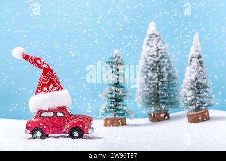 Macchinina giocattolo rossa con un cappello di Babbo Natale sul tetto accanto agli abeti mentre nevica. Concetto invernale e natalizio. Foto Stock