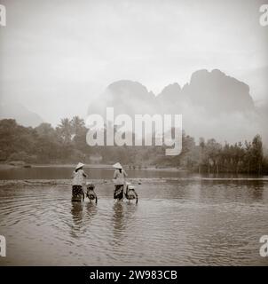 Due donne che indossano conica cappelli asiatici spingere le biciclette attraverso un fiume poco profondo in Vang Vien, Laos. Foto Stock