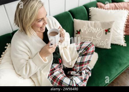 Donna che tiene una cioccolata calda seduta sul divano e indossa abiti comodi Foto Stock
