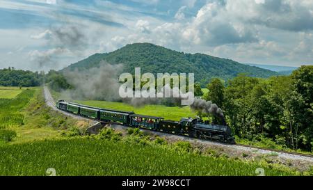 Una vista aerea di un treno passeggeri a vapore a scartamento ridotto, che si avvicina viaggiando intorno a una curva attraverso la campagna, soffiando fumo, in una soleggiata giornata estiva Foto Stock